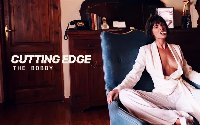 AI-generated image: The Bobby, The Cutting Edge: a woman with a dark layered French bob ending at the mouth, with a short fringe, in an ivory suit, leaning back in a blue velvet armchair with a cigarette between her teeth, in a wood-panelled room