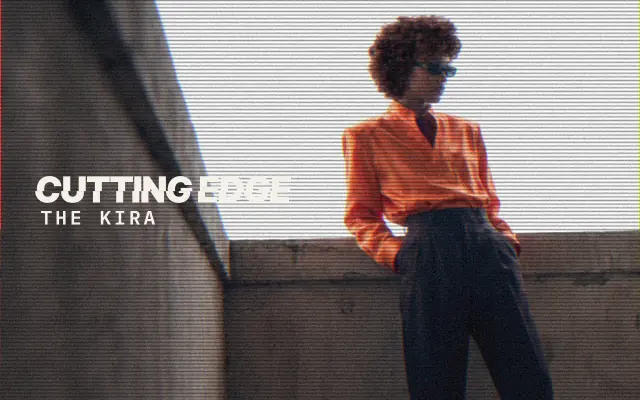 AI-generated image: The Kira, The Cutting Edge: a woman with a short rounded curly cut and a curly fringe, in an orange satin shirt and sunglasses, leaning on a concrete ledge