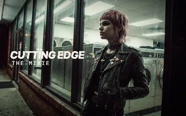 AI-generated image: The Mixie, The Cutting Edge: a woman with a near-black mullet pixie, short choppy fringe and wispy nape pieces, in a black bomber and silver chains, leaning in beside the washers of a late-night laundromat