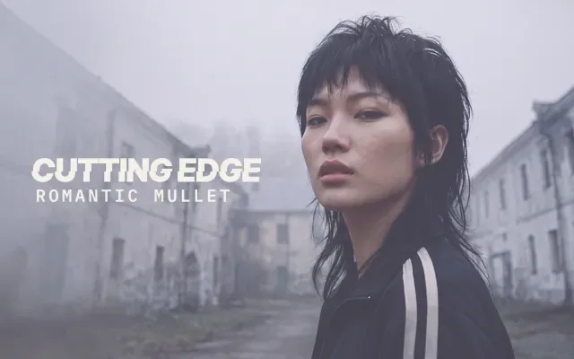 AI-generated image: Romantic Mullet, The Cutting Edge: a woman with a black shaggy mullet and choppy fringe in a black track jacket, looking back over her shoulder in a foggy courtyard of old grey buildings